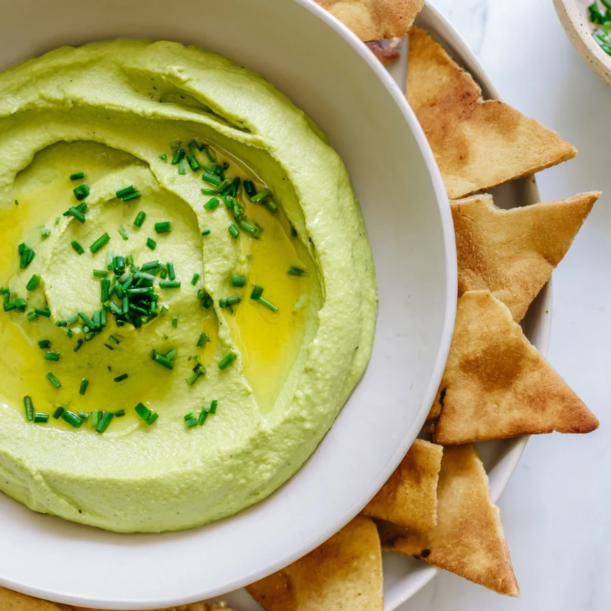 Smooth, herb-flecked green avocado hummus garnished with olive oil and fresh cilantro, beside crispy pita triangles.