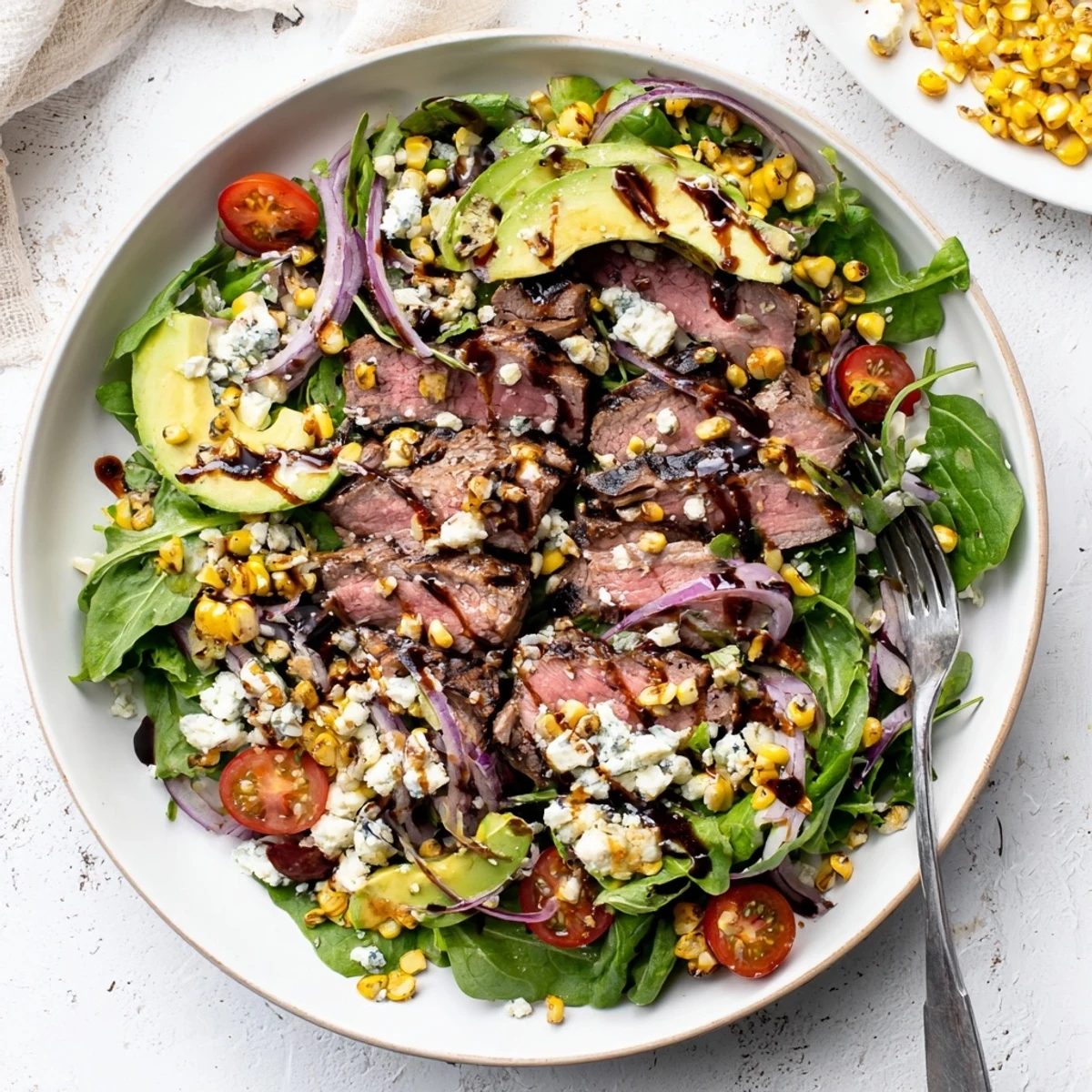 A vibrant balsamic steak Gorgonzola salad with grilled corn topped with juicy sliced steak, creamy cheese, and sweet corn on a white platter