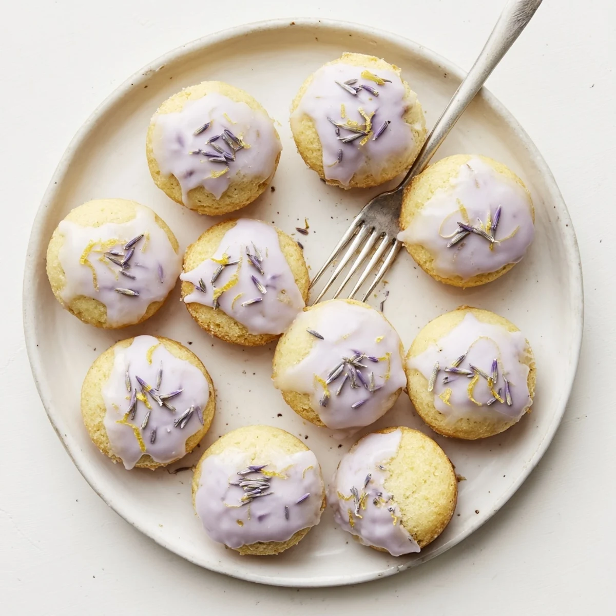 A batch of twelve mini lemon cakes topped with glossy lavender glaze and lemon zest