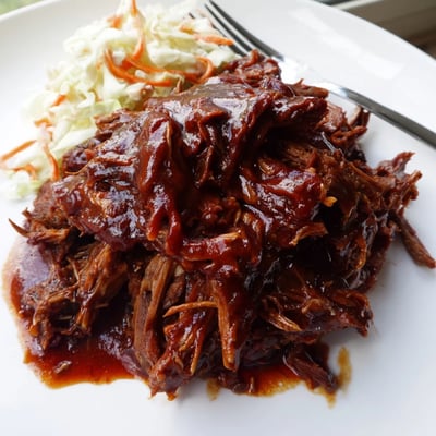 Tender slow cooker BBQ pulled beef glistening with tangy sweet sauce on a rustic plate